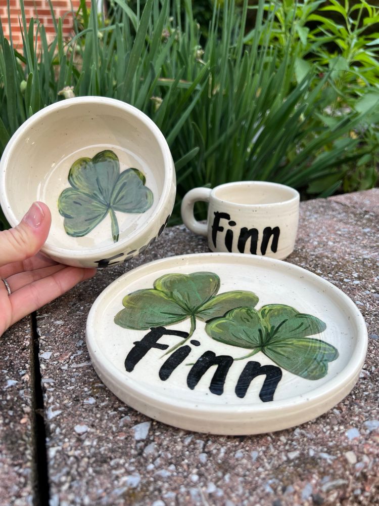 Handmade wheel thrown stoneware pottery set by kate rowe featuring a plate, a bowl, and a small mug all with hand painted shamrocks and the name Finn in black lettering