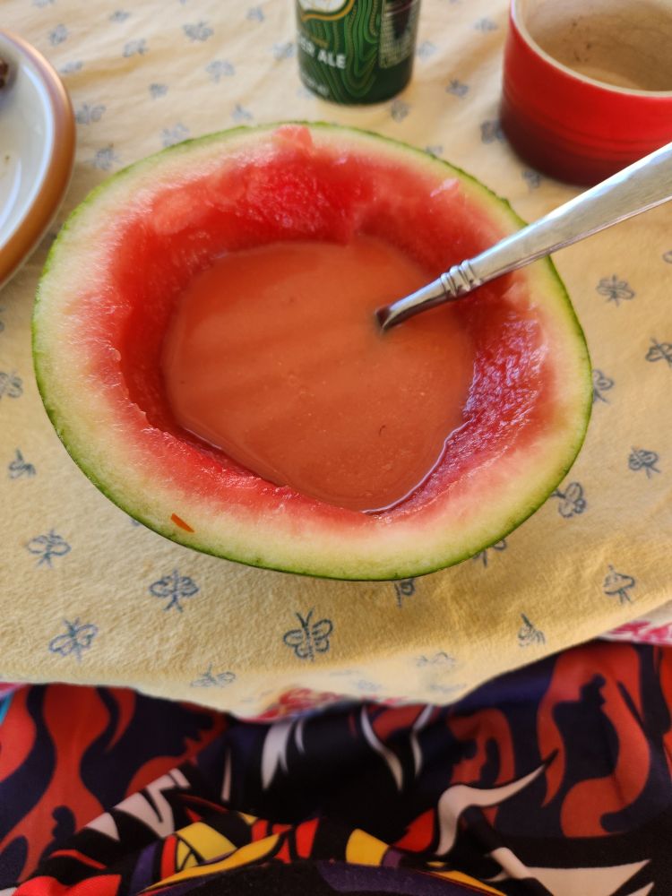 A small, hollowed-out watermelon half filled with pink soup. A spoon is sticking out of it