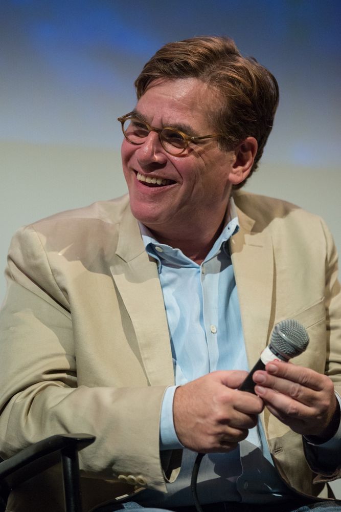 Aaron Sorkin, wearing a tan suit jacket, blue button down shirt, and holding a microphone while sitting. He is smiling and wearing glasses.