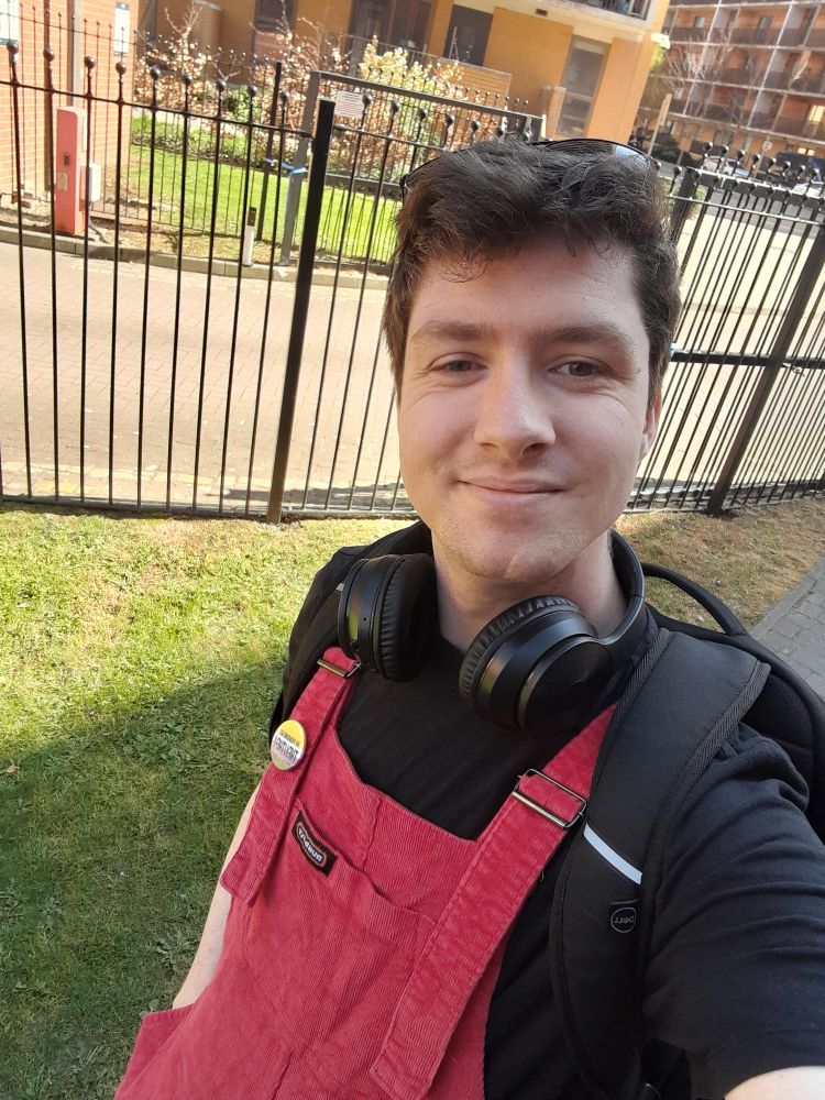 A selfie of Matt walking outside. They are wearing red dungarees, headphones, and sunglasses on their head.