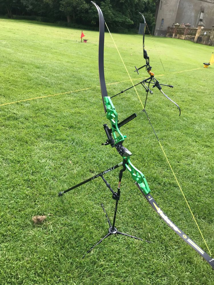 Picture of a green bow with neon yellow string, black limbs with a black sight and black long rod attached, resting on a black stand. The grass is green and in the background is another bow but black with an orange grip.