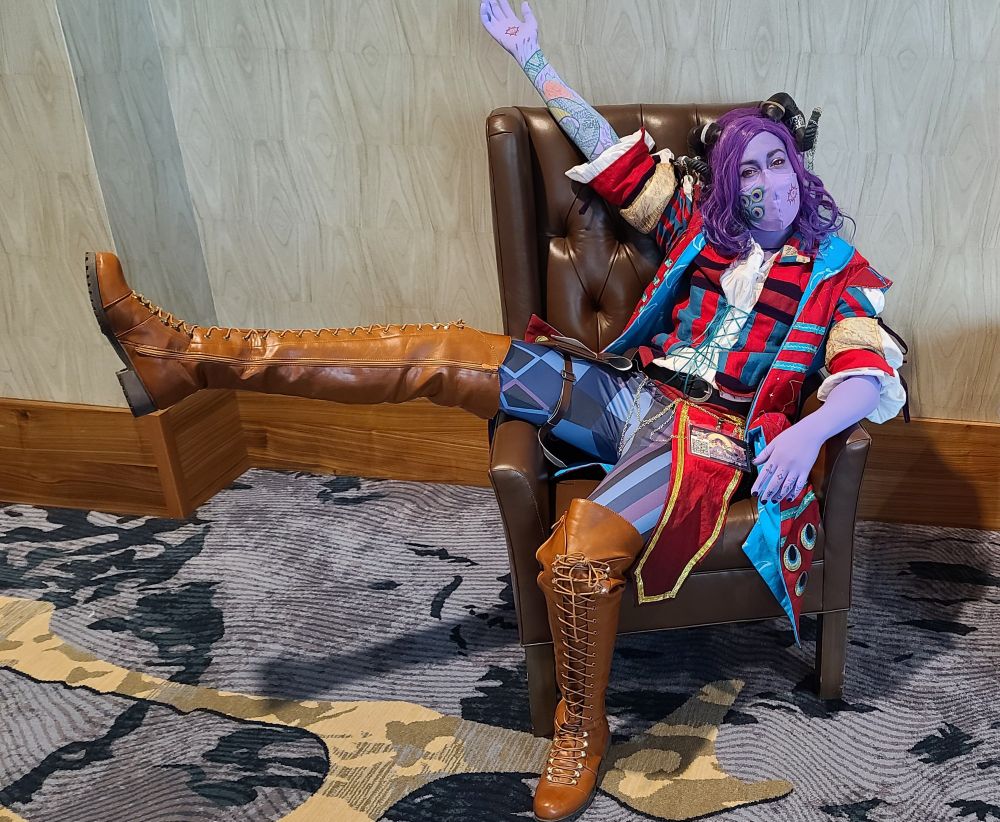 A person cosplaying as Molymauk Tealeaf from the Dungeons and Dragons podcast, Critical Role. They are relining, lopsidedly, on a leather chair in a hotel hallway. One arm is held aloft, and one leg is sticking out over the armrest. The person is wearing lavender facepaint, and long laveder gloves to depict the character's purple skin. One glove has some tattoos drawn on. The person is also wearing a purple face mask with some tattoos drawn on. The person has thigh high leather boots, purple and blue patterned leggings, a colorful shirt, a long colorful jacket, a wavy shoulder length purple wig, and some curled horns adorned with chains.
