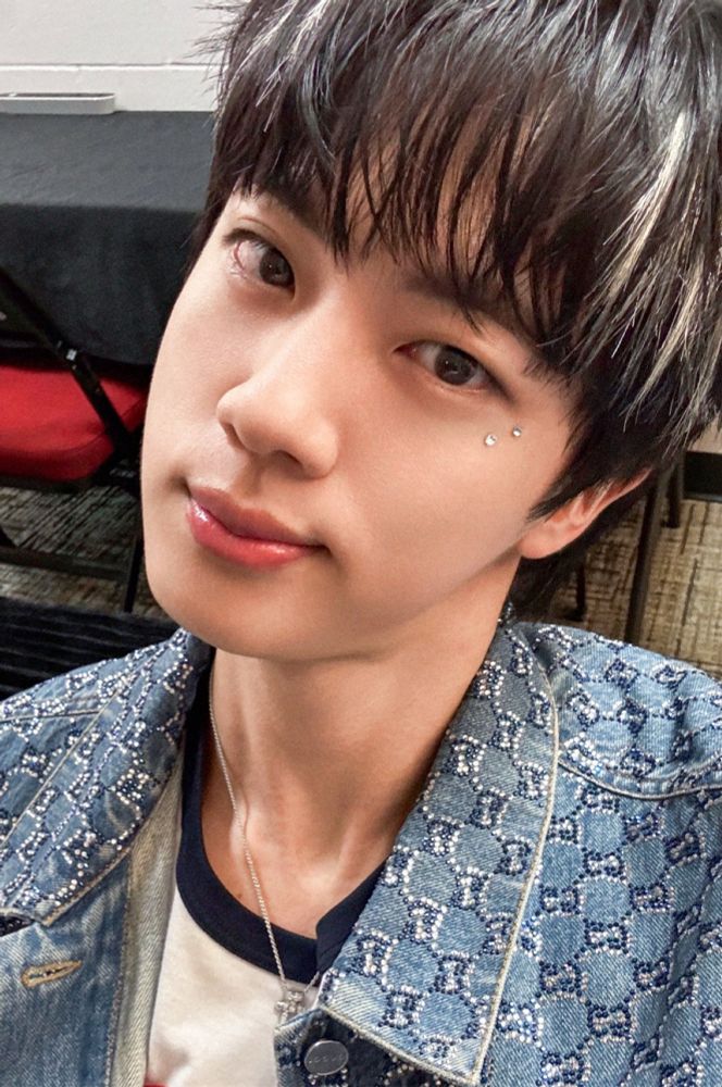 Jin smiling with head tilted, blonde highlights and gemstones by his eye 