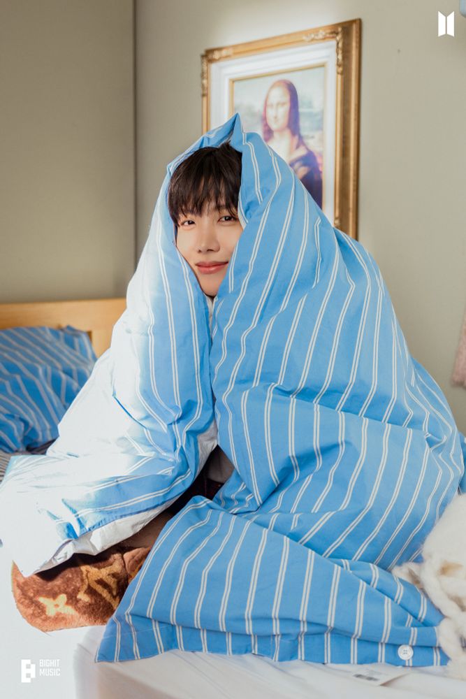 Hobi sitting on a bed wrapped in a puffy blue comforter 