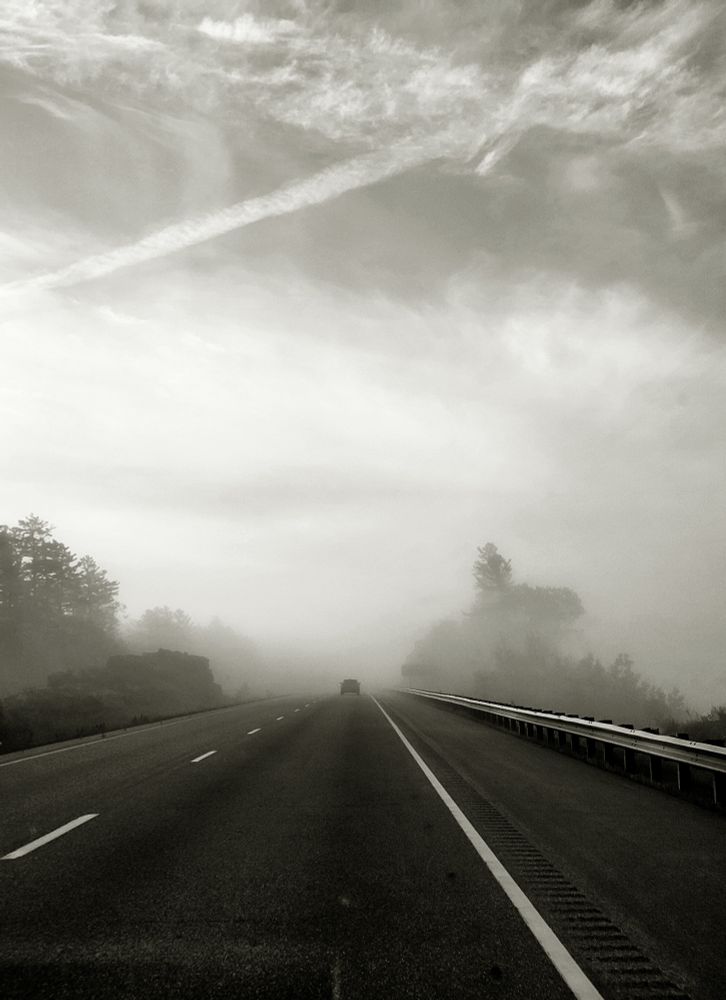 A black and white image of the painted lines on a highway and the railing beside the asphalt as they stretch toward a distant, barely-visible vehicle on a foggy day. 