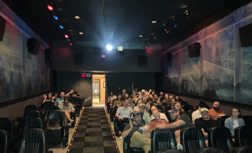 A movie theater auditorium, photographed from the screen with the projector in the background and a crowd of people filling most of the seats. They look excited. 