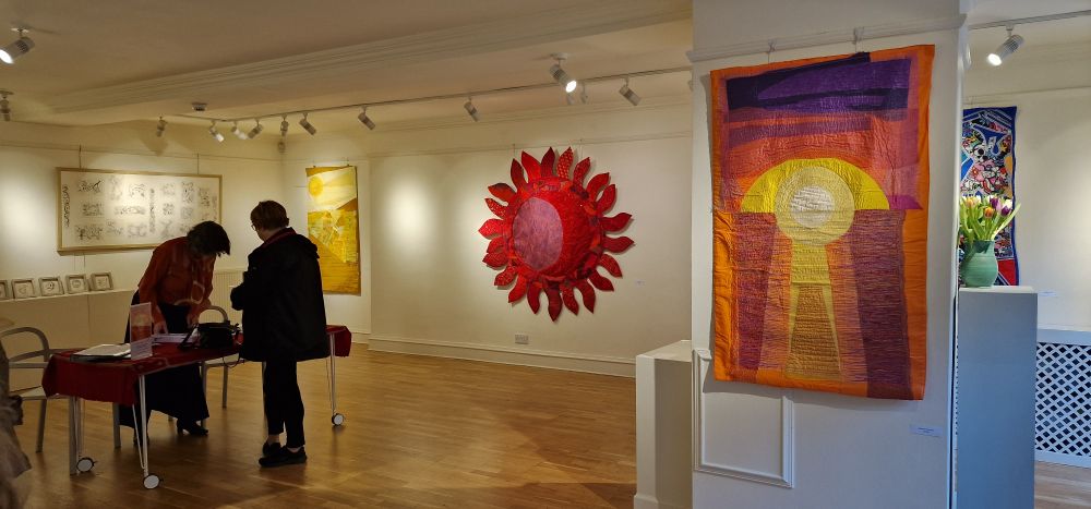 A well lit, low cielinged art gallery with light cream walls. On the walls hang brightly coloured textile art pieces depicting fire, suns, rolling hills and water. Two people stand off to the side, chatting over a welcome table. 