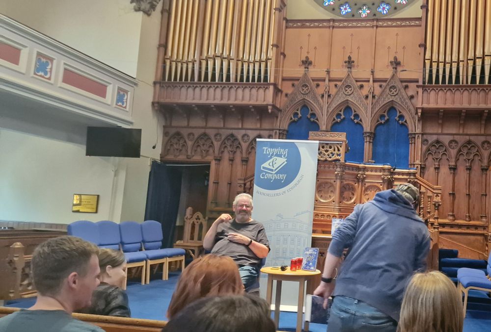 Ben Aaronovitch, author of the Rivers of London series, sits on a chair in a big church auditorium looking at an audience of fans. Everyone is smiling and he has his new book 'Stone & Sky' sitting by his side, ready for reference in questions and answers. 