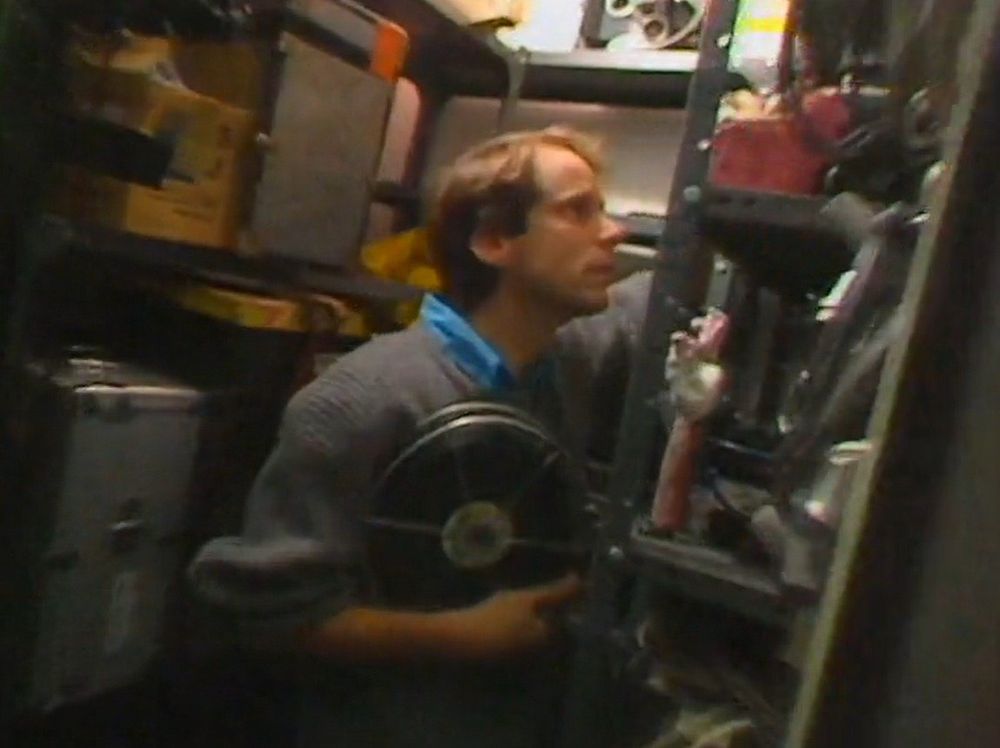 Still from a documentary on animation, featuring a young man (Nick Park) standing in a cramped storage cupboard, holding a film reel. On a shelf in the cupboard, there is the model of his character, Wallace.