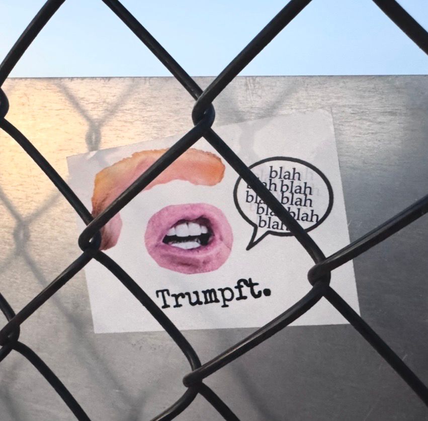 A sticker behind a chain-link fence has a cut out picture of Donald‘s hair and mouth and a word bubble that says “blah blah blah, blah, blah, blah, blah blah“

It reads:

Trumpft.