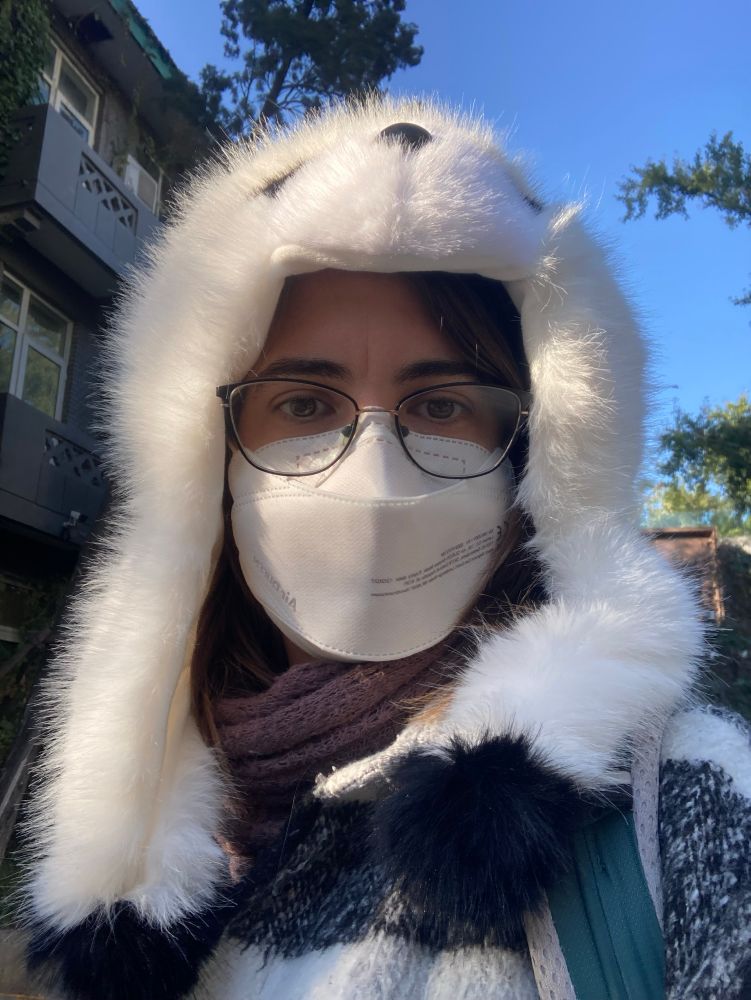 Selfie de uma mulher branca de óculos de grau e máscara com um gorro branco mitando a cabeça de um panda com abas pra proteger as orelhas