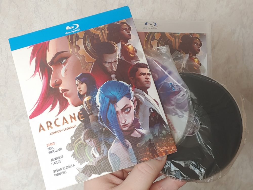 Me holding the Arcane Blu-ray set kinda awkwardly. There's a slipcover, disc case, and two discs visible. The case and slipcover have the exact same art showing the main cast of characters from the show.