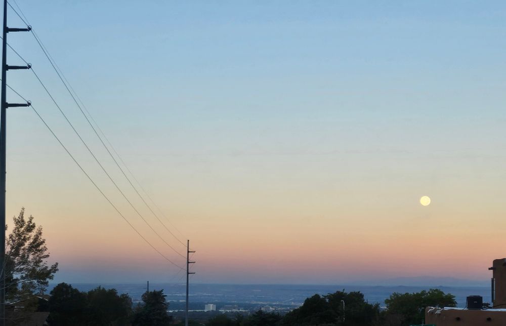 Full moon on the westward horizon and a view overlooking Albuquerque. The morning sky is filled with dawn colors, rose pink, orange yellowing into pale blue and purple while the city wakes up
