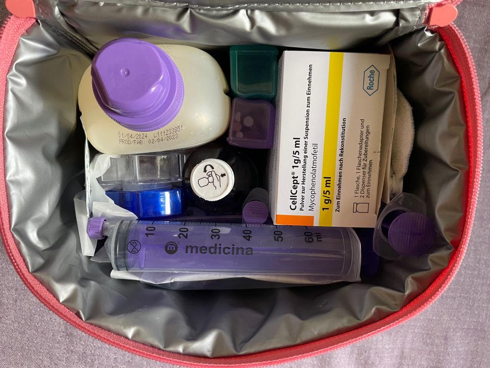 Blick von oben auf eine geöffnete Thermostasche. Innen Silber/grau, am Rand Pink. Darin erkennbar mehrere Sondenspritzen, eine Medikamentenflasche, 2 Behälter mit Tabletten in lila und türkis, eine Packung Cellcept Lösung, ein Tablettenmörser und eine Flasche Sondennahrung.