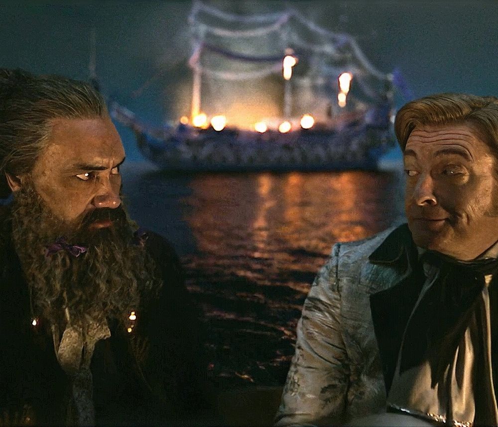 A scene from s1. Ed and Stede are in a dingy. Ed is looking at Stede with intensity and Stede smiles at him while the French party boat burns behind them. 