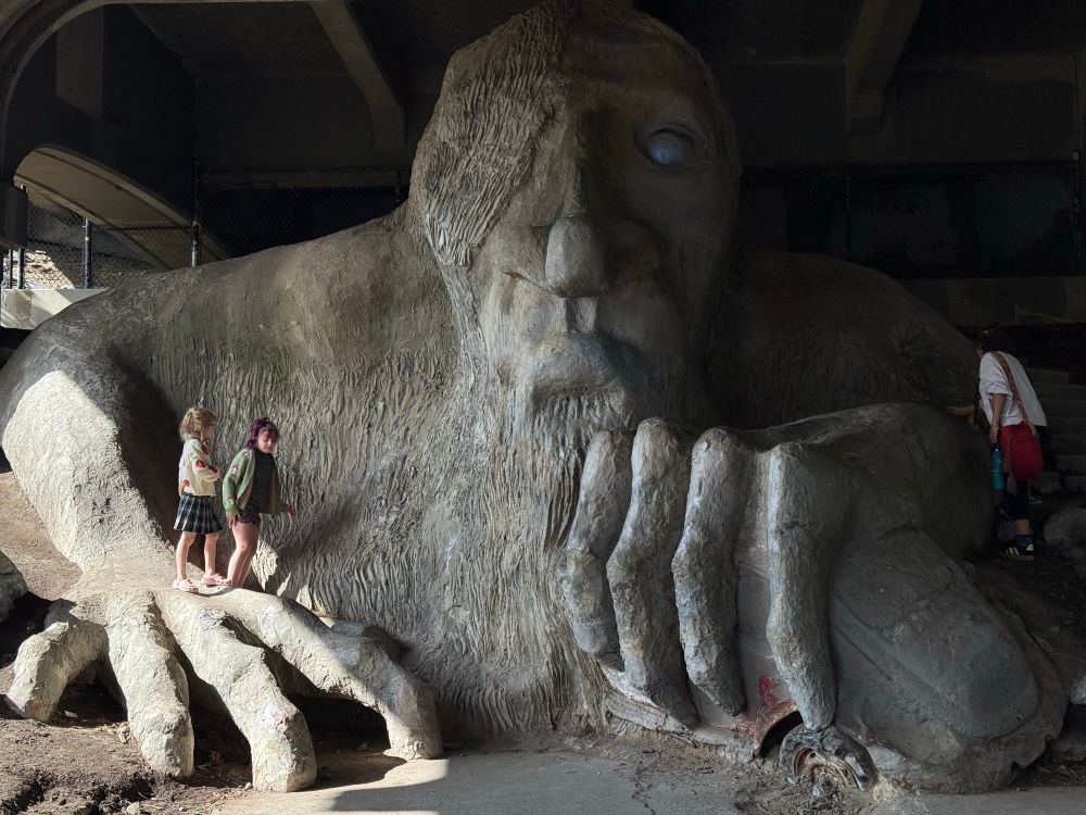 A few children and tourists climb on the Fremont Troll, a huge sculpture that lives under a bridge in the Fremont neighborhood of Seattle Washington, USA. The troll holds an actual concrete-coated VW beetle.