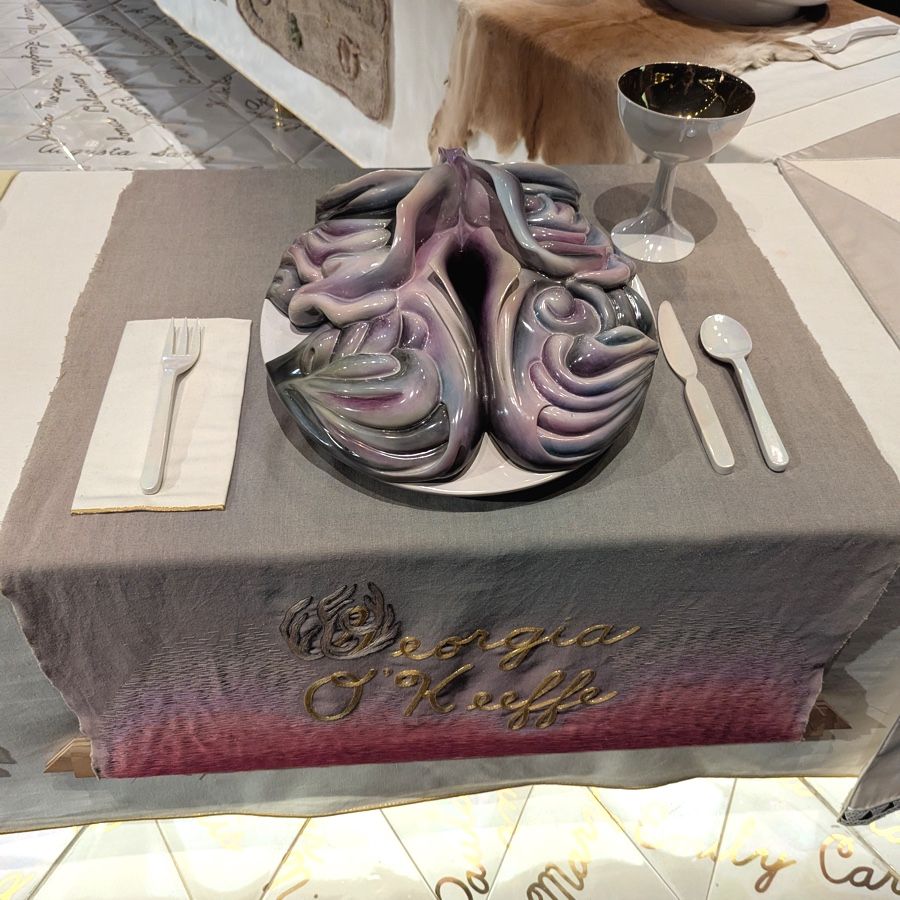 Photo of a table setting. The runner under the plate is gray, and has the name "Georgia O'Keefe embroidered in gold script. The place setting consists of a plate, napkin, fork, spoon, knife, and goblet. The plate has been elabroated with a complex, organic-looking multi-layered sculpture." 