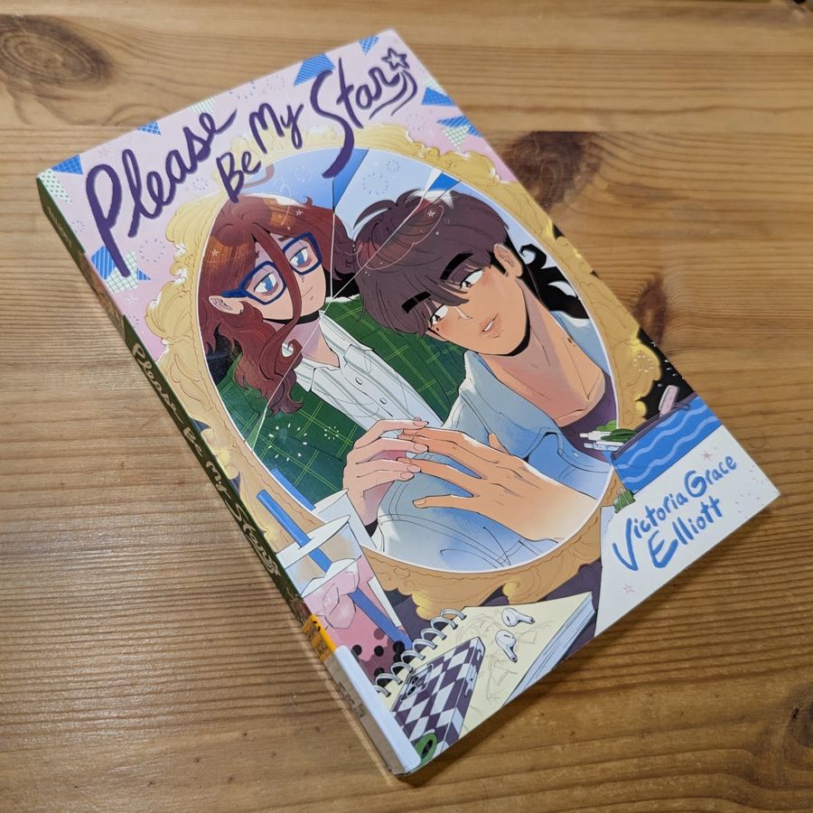 Photo of a softcover book on a wood surface. Title is PLEASE BE MY STAR in dark purple hand-drawn script. Illustration shows an oval-shaped mirror, cracked, reflecting a young man and woman; she's behind him, reaching her hand over his shoulder, and he's reaching his hand back, their figertips just touching, Next to the mirror are a cellphone and some earbuds sitting on a drawing pad, a clear cup of boba tea, and a pen case with some pens in it. Author's name is Victoria Grace Elliott. .