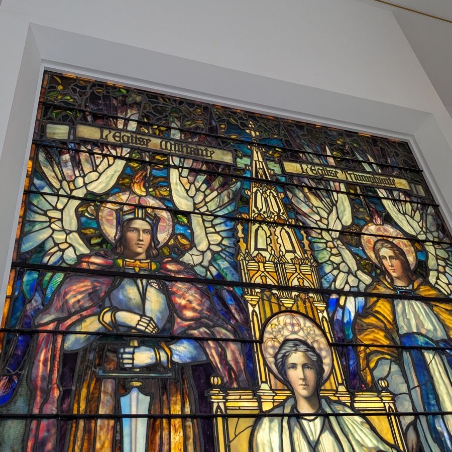 Photo of part of a stained-glass window, showing angels with swords flanking a church steeple and a saint. (Not sure which saint.) Banners at the top say "L'Eglise Militante" and "L'Eglise Triumphante."