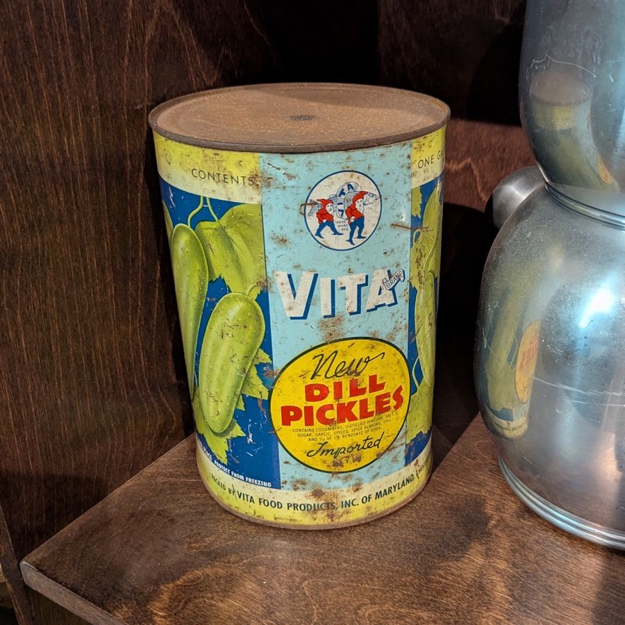 Photo of a canister of Vita brand dill pickles on a shelf. The canister looks like it was designed in the 1960s or '70s, and is battered and stained. 