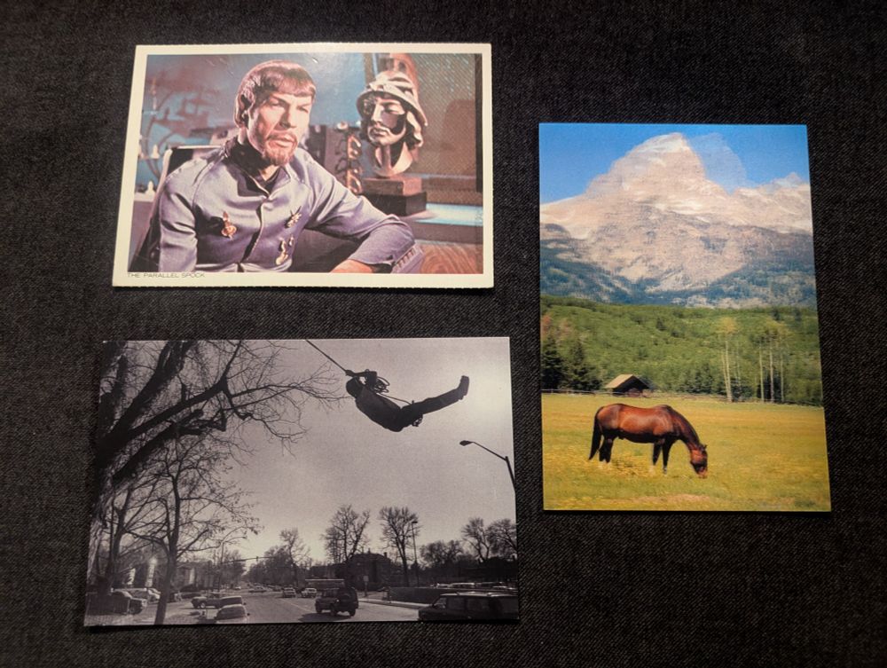 Three postcards on a dark blue fabric background. The top left is of the evil mirror universe version of Spock from Star Trek. The bottom right is a black and white photo of a man using a rope swing over a street. The right is a scene of a horse grazing in a field in front of the mountains of Jackson Hole, Wyoming.