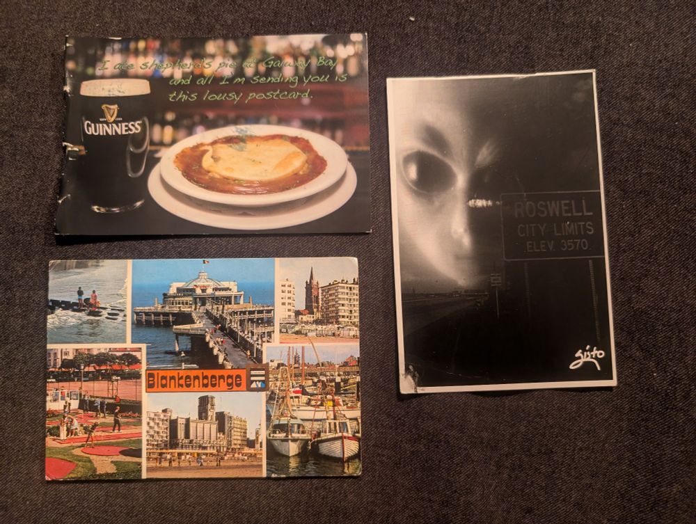 Three postcards on a gray fabric background. 

The card in the top left shows a picture of a pint of Guinness and a plate of shepherd's pie on the counter at a pub. The green text on it reads "I ate Shepard's pie at Galeway Bay and all I'm sending you is this lousy postcard."

The bottom left postcard is from the city of Blankenberge, Belgium. It is a multiview card with six pictures and the text "Blankenberge" in black font on an orange background. The pictures on the card (clockwise from the top left) are of people standing on a submerged bridge in the water, a large building on the end of a pier, a collection of buildings including a Church steeple, boats in a marina, a view of buildings from the beach, and several groups of people playing at a putt-putt golf course.

On the right, the postcard shows the semi-transparent face of a gray alien over a picture of the outskirts of Roswell, New Mexico where there's a sign that reads "Roswell City Limits Elev 3570."