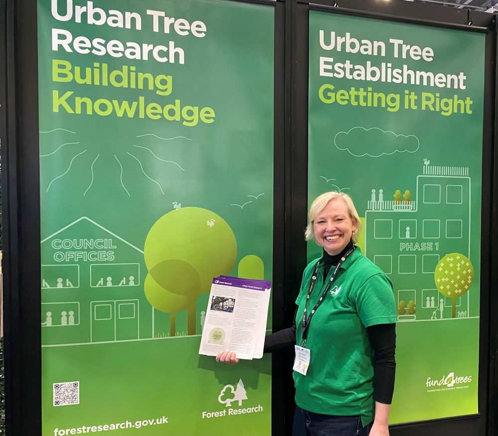 Claire Woodall stands in front of a green display holding a publication. The display reads "Urban Tree Research - Building Knowledge".
