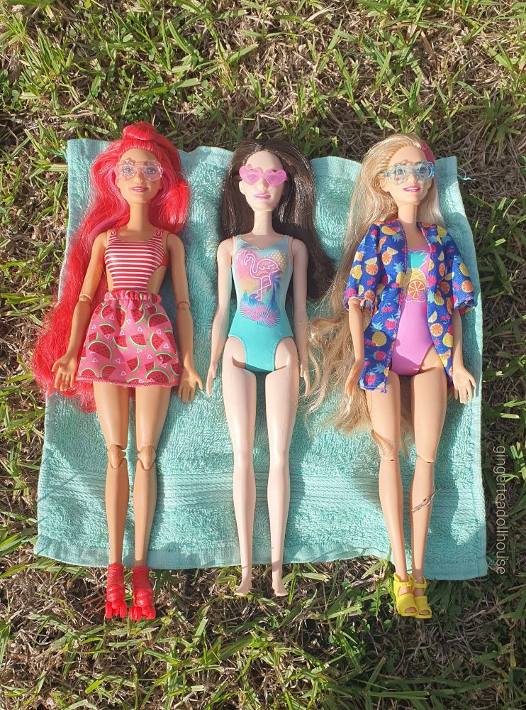 From left to right: Barbie Pop Reveal Fruit Series Watermelon Crush, Beach Barbie and Barbie Pop Reveal Rise and Surprise laying down on a towel trying to catch some sun rays!