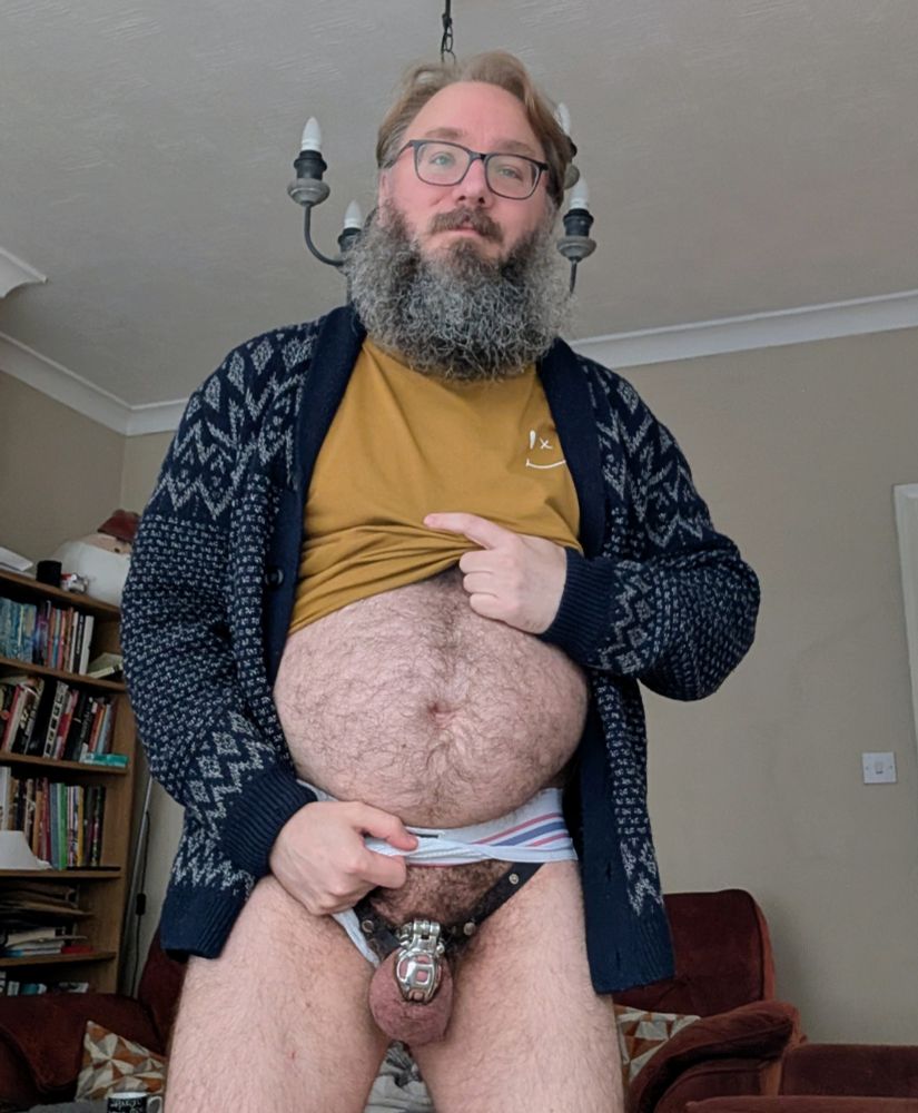Wearing a navy cardigan and brown t shirt, and just a white jock below, pulled aside to show chastity caged nub 