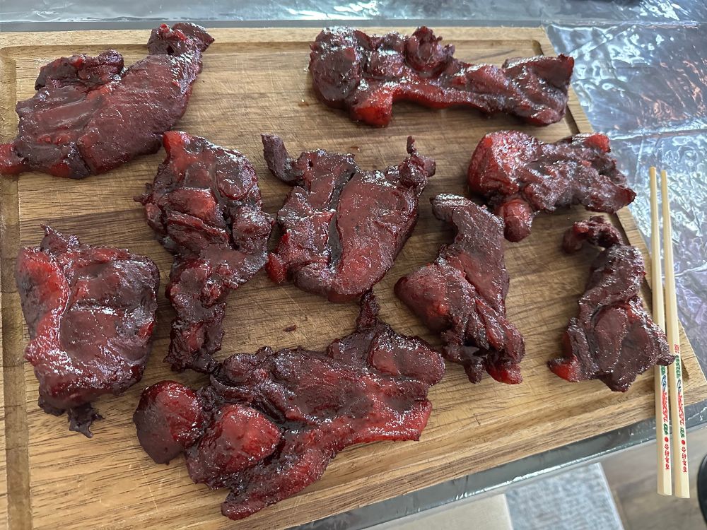 Finished char siu, gleaming dark red and brown, sitting atop a brown cutting board with a pair of chopsticks. The board is on a plastic transparent table cloth on a brown table.