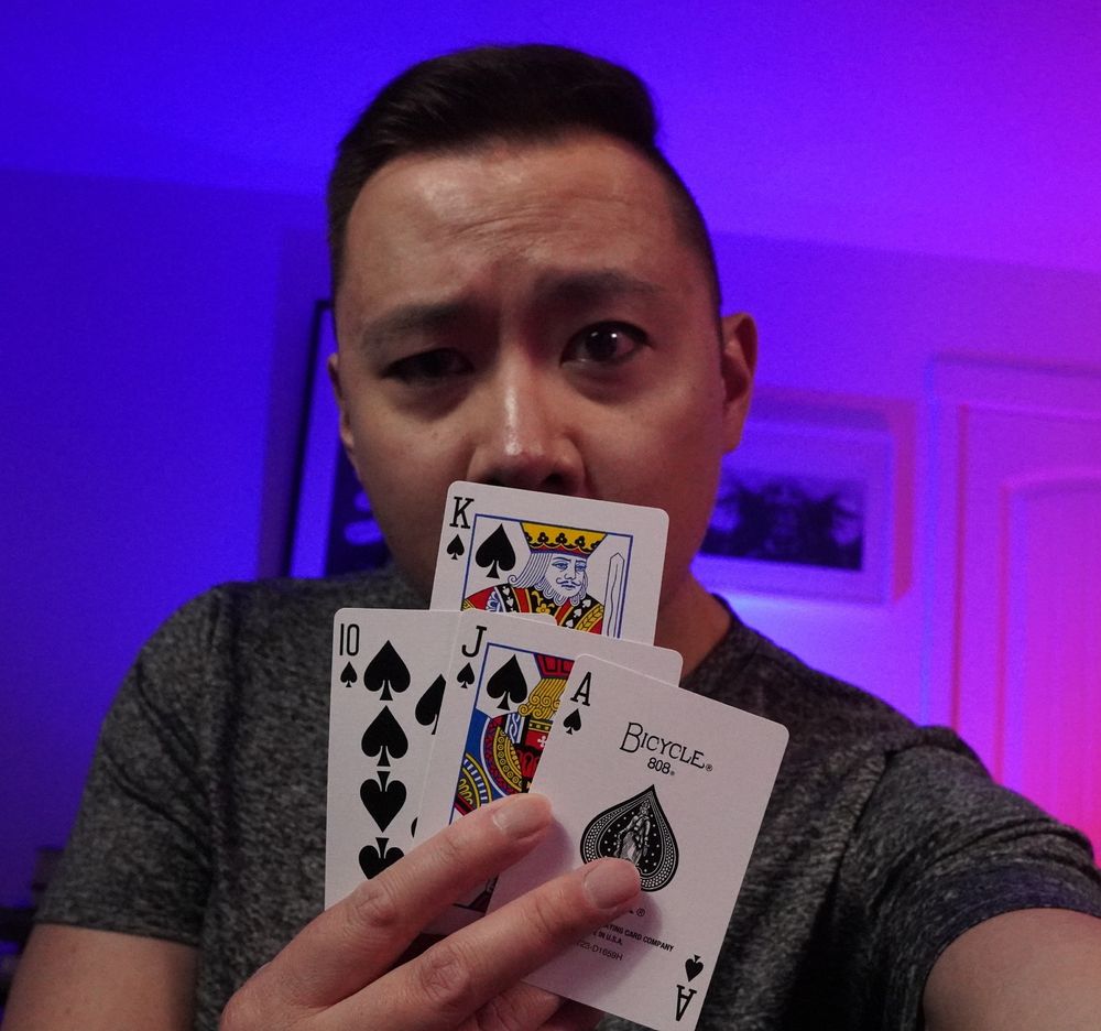 Matt is wearing a grey shirt, giving a shifty eyed look at the camera whilst partially hidden behind a 10, Jack, King and Ace of Spades