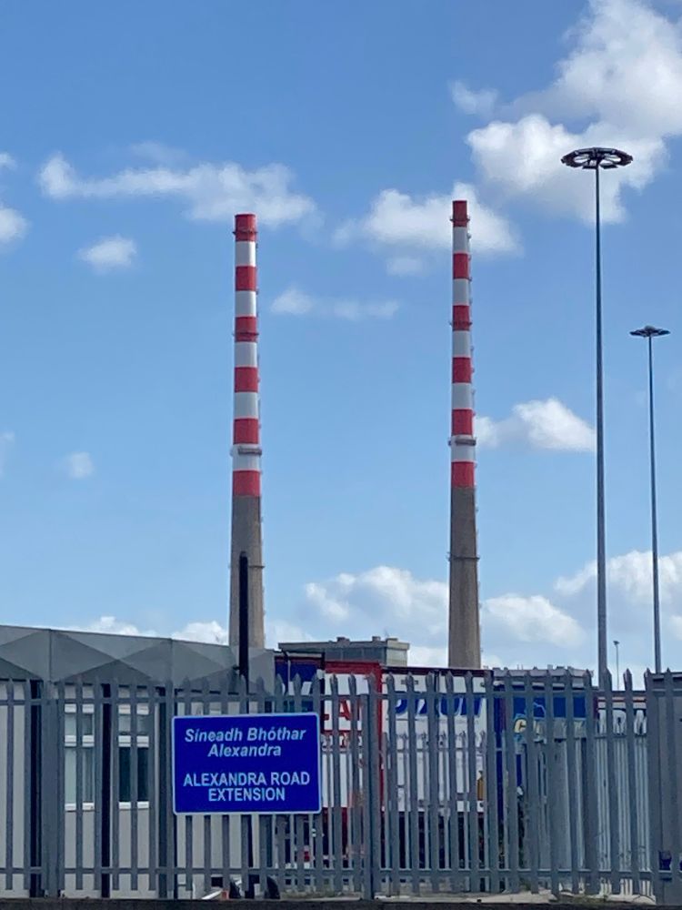 Two red and white chimneys against a pale blue sky. There are industrial buildings below them and a grey steel fence in front with a big blue sign on it that reads Alexandra road in white letters