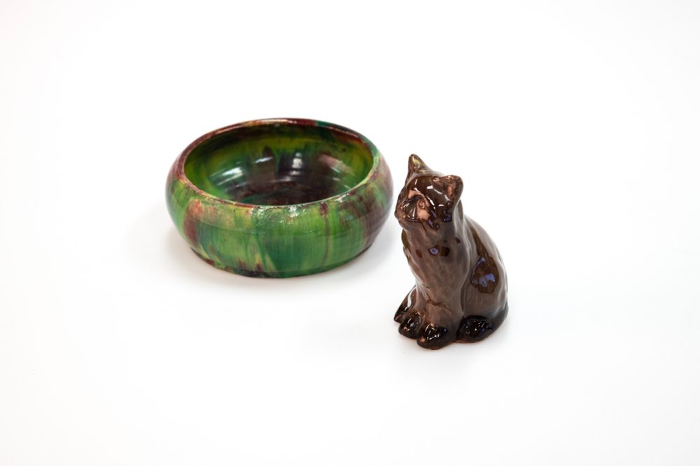 A glossy, dark brown ceramic cat figurine sits next to a shallow bowl with a vibrant green, red, and brown mottled glaze. Both objects are positioned on a solid white background.