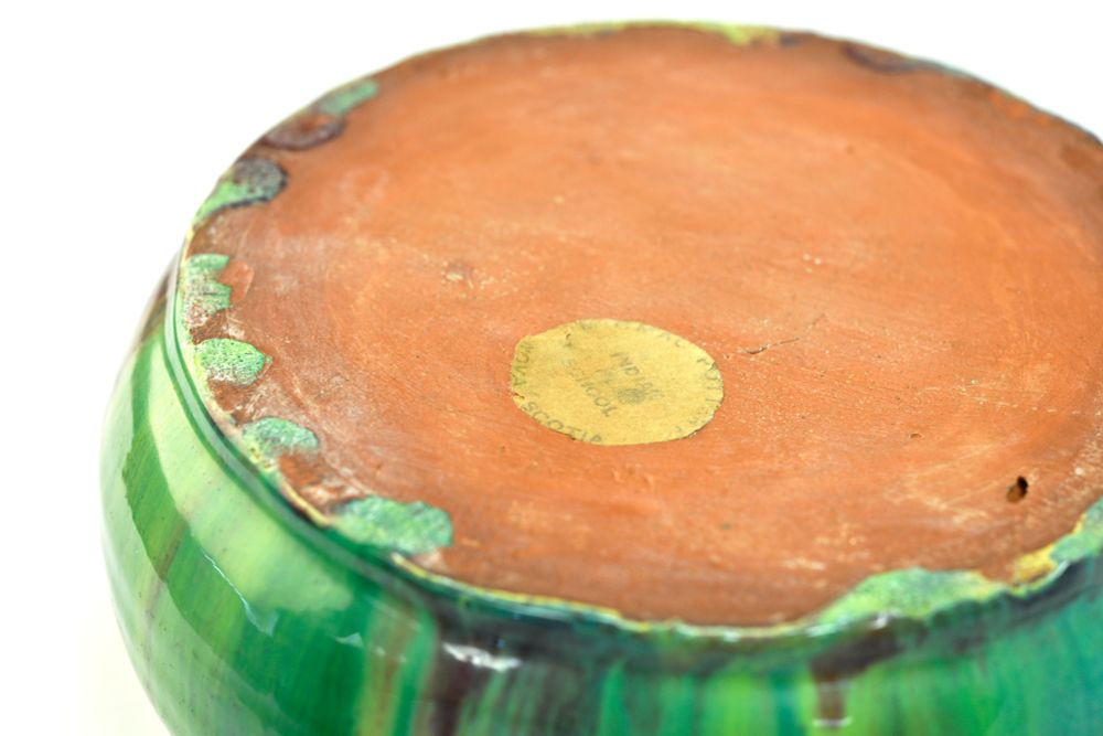 The unglazed, orange-brown ceramic base of a bowl with a colorful green, red, and brown mottled glaze visible around the bottom rim. A small, circular, faded yellow label is affixed to the center of the terracotta base, reading 'INDIAN RES. SCHOOL SHUBIE NOVA SCOTIA,' referring to the Shubenacadie Residential School.