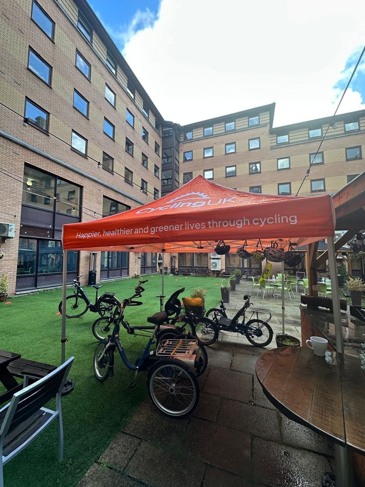 Orange Cycling UK tent set up in a courtyard with several adaptive bicycles parked underneath on artificial grass surrounded by multi-story brick buildings.