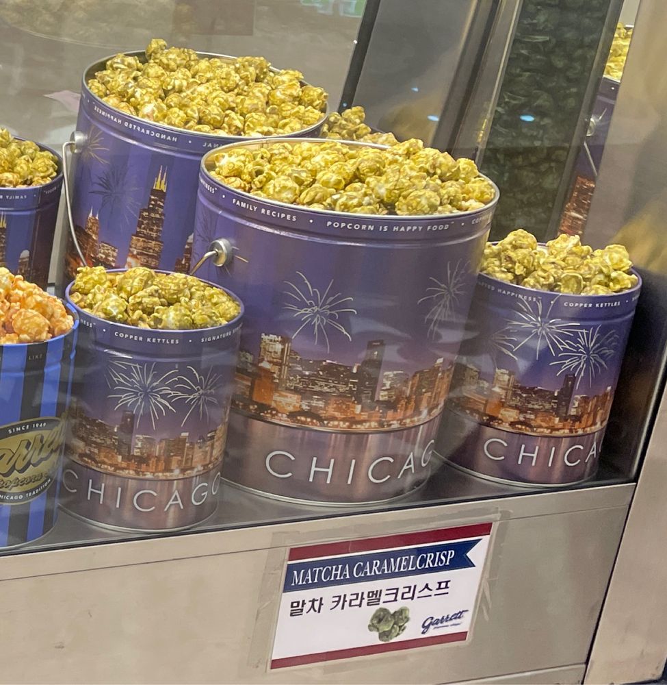 Various sizes of tins brimming with green-tinted popcorn, with a label reading "Matcha Caramel Crisp 말차 카라맬크리스프"