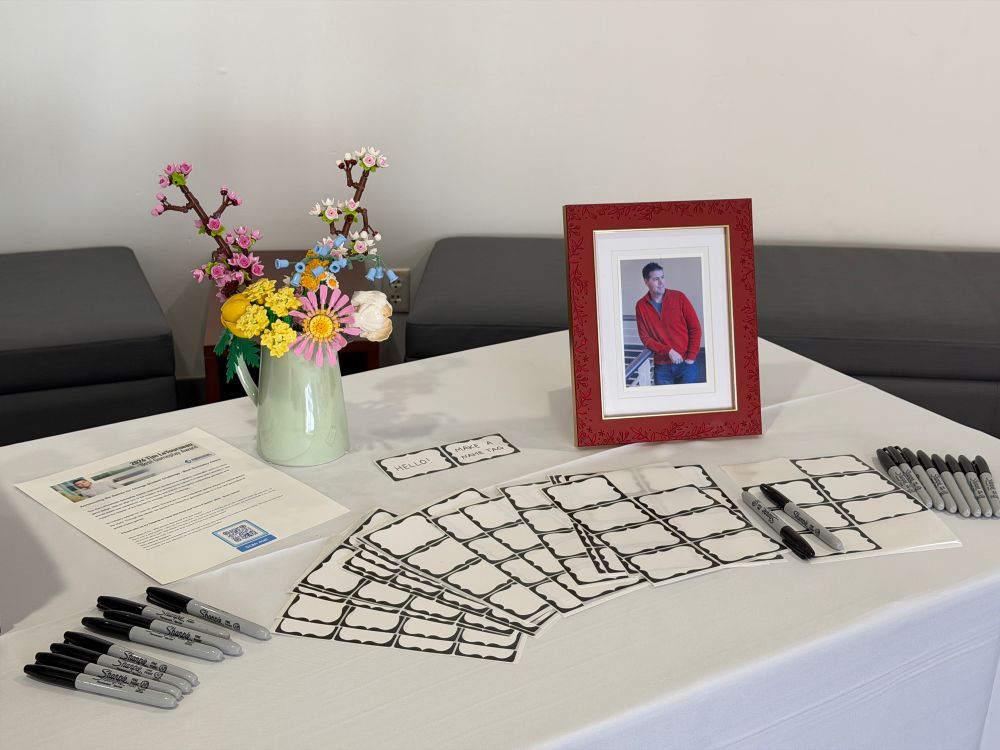 The right end of a guest reception table, featuring a handsome framed photo of Tim LeTourneau in his trademark red zipper sweater, blank name tags with Sharpie permanent markers, and a Lego flower bouquet in vase.