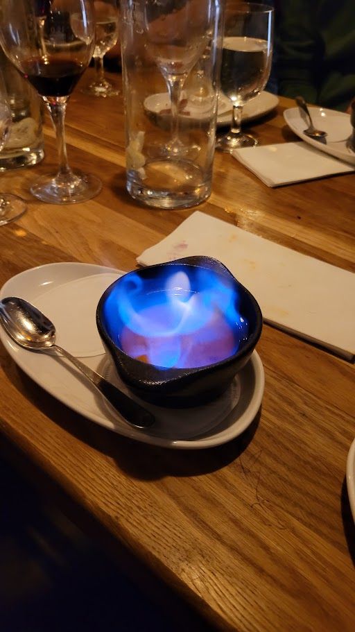 Eine kleine, schwarze gußeiserne Schale auf einer ovalen, weißen Untertasse mit Löffel. In der Schalde brennt mit blauer Flamme der Alkohol. 