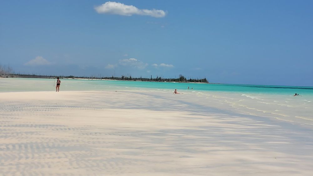 Im Vordergrund weißer Strand, feucht vom Wasser. Auf mittlerer Distanz einige Leute, die im Wasser baden. Im Hintergrund etwas tieferes Wasser, das hell-türkis ist. 