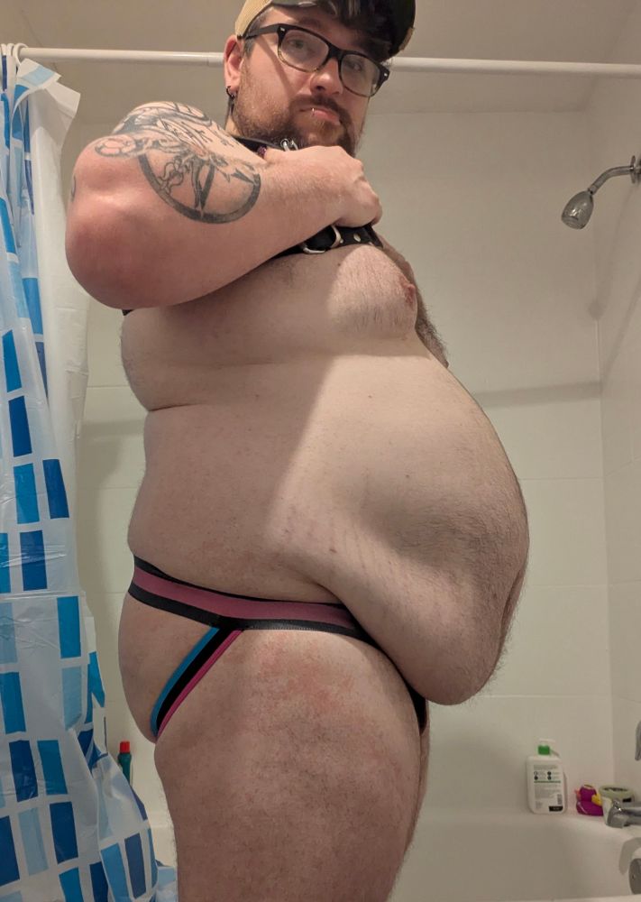 Me in a harness, hat, jockstrap, and glasses in my bathroom.