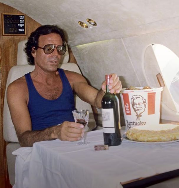 Julio Iglesias en lunettes de soleil aviateur dans son jet privé, avec une bouteille et un verre de rouge accompagné d'un bucket de tenders KFC 