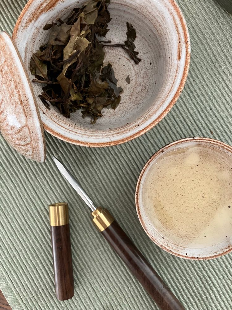 steeped tea leaves in a gaiwan next to a small teacup with light yellow tea and a woof and metal pick used to pull the tea cake apart 