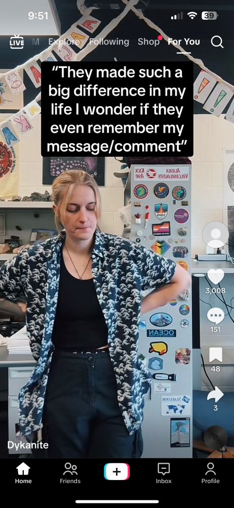 A young and cool geologist in a patterned button down saying goodbye to TikTok from inside their lab. Behind them is gray lab equipment with color magnets and stickers.
