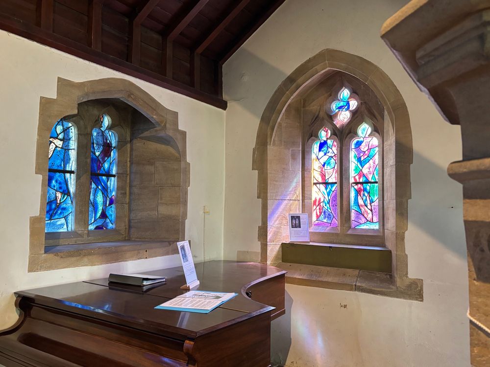 Two modernist stained glass windows in a church, with a grand piano in front. One window is mainly blue and the other also has reds and purples, the sun is shining through creating a purple and orange reflection on the stone wall. The windows are designed by the artist Marc Chagall. 