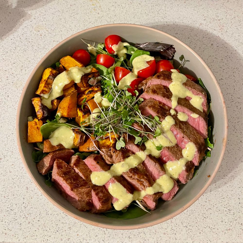 Bowl with grilled steak, roasted sweet potatoes, spinach, cherry tomatoes, finished with a creamy pesto sauce and micro greens 