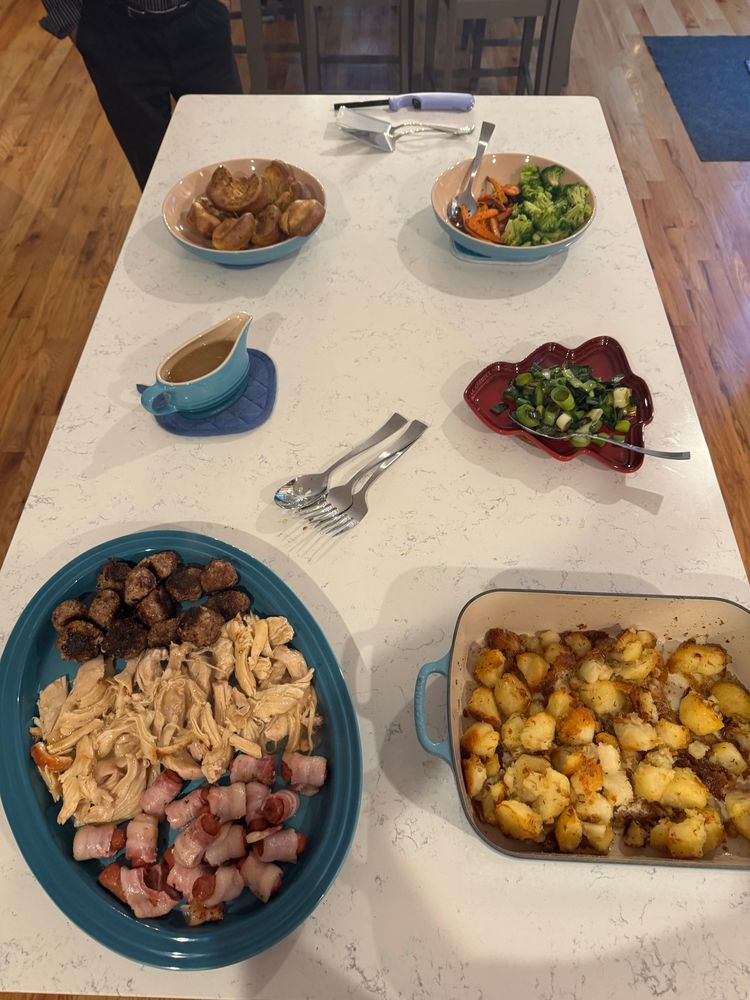 An arrangement of food on platters. Turkey, stuffing and pigs in blankets are on a turquoise plate. Roast potatoes, leeks, broccoli, carrots, gravy and Yorkshire puddings are also on the kitchen island. 