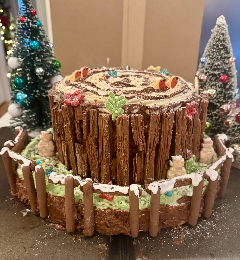 A cake shaped like a tree trunk. The top of the cake is cream butter icing with melting chocolate in rings. The edge is chocolate butter icing with Flakes stuck in. The base is green icing and there is a fence made of chocolate finger biscuits on the front with white icing for snow on top. Around the cake are decorative icing shapes - robins, mistletoe, and poinsettias. There is also 3 peppermint bark snowmen standing next to the tree trunk. In the background there are 3 mini fake Christmas trees. 