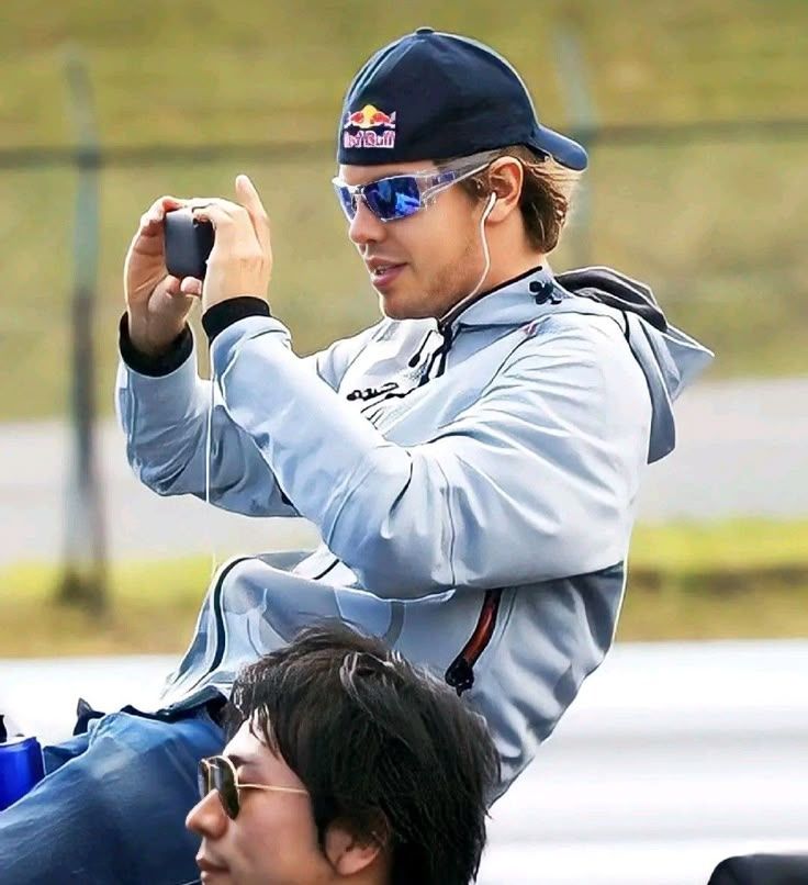 sebastian vettel circa probably 2009ish. he's got a red bull cap on and the most early 2000s clear sunglasses possible. he's wearing wired earphones and plugged into his phone, which he's using to take a picture on. 