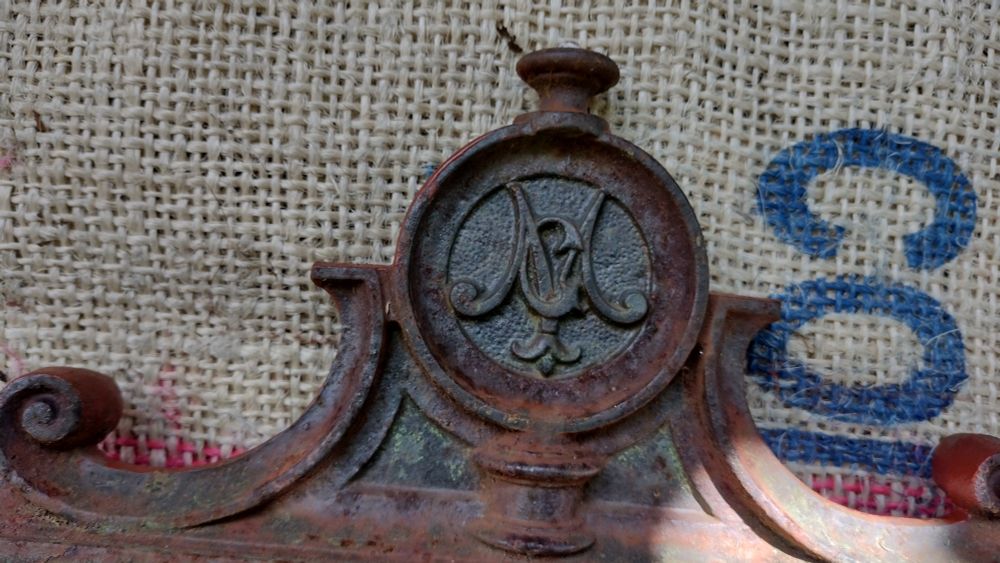 Part of a very rusty mantle showing an emblem with the Letters M and G cast into the iron.  It has been painted with red oxide primer 