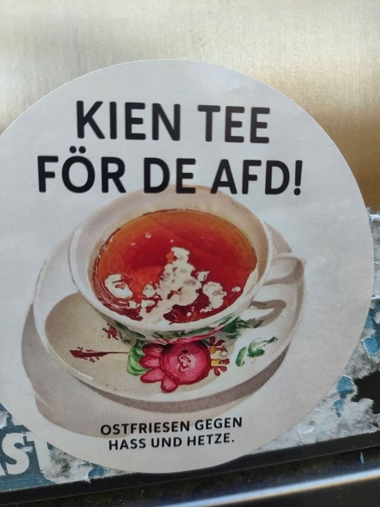Zu sehen ist ein Runder Sticker mit einer mit Blumen bemalten nostalgisch wirkenden Porzellantasse mit schwarzem Tee und Milchschlieren darin. Über der Tasse steht "KIEN TEE FÖR DE AFD!". Unter der Tasse steht: "OSTFRIESEN GEGEN HASS UND HETZE."
